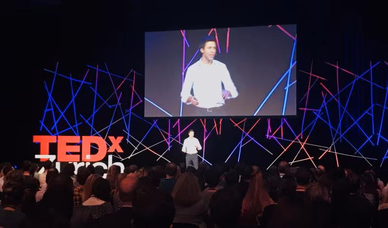 A speaker on the TEDxZurich 2018 stage, framed by a web of glowing blue and red light lines
